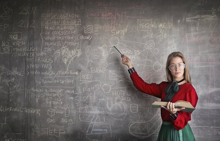 serious female teacher wearing old fashioned dress and eyeglasses standing with book while pointing at chalkboard with schemes and looking at camera 3771074 scaled